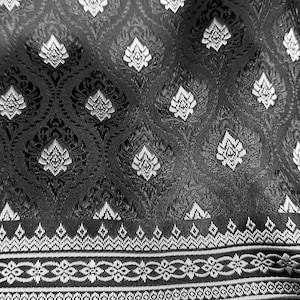 May include: Close-up of a black and white patterned fabric. The design features a repeating floral motif across the main body, with a detailed border of geometric and floral patterns. The fabric has a woven texture.
