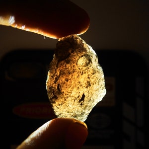 Pode incluir: Uma pedra translúcida, de forma irregular, cor âmbar, é segurada entre dois dedos. A pedra tem uma superfície áspera e texturizada com pequenas reentrâncias. A luz brilha através da pedra, realçando suas características únicas.