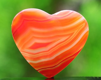 Puede incluir: Una piedra de ágata en forma de corazón con rayas naranjas y blancas. La piedra está pulida y lisa.