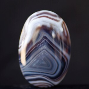 Puede incluir: Primer plano de una piedra de ágata con bandas blancas, marrones y grises. La piedra tiene una superficie lisa y pulida y tiene forma ovalada.