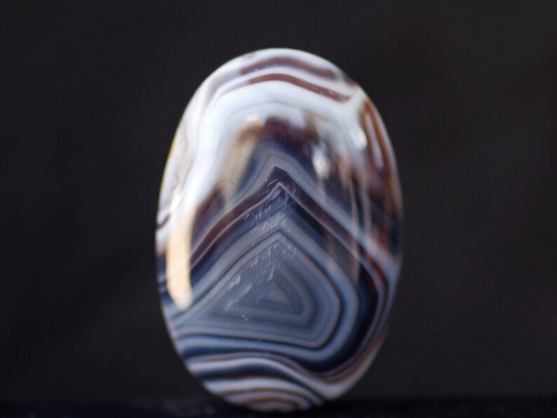 May include: A close-up of a white, brown, and gray banded agate stone. The stone has a smooth, polished surface and is oval-shaped.