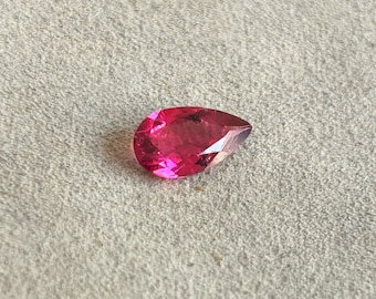 May include: A teardrop-shaped, faceted gemstone with a deep pink hue. The gemstone is set against a light gray background.