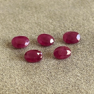 Könnte beinhalten: Fünf lose, oval geformte Rubine mit Facettenschliff. Jeder Edelstein zeigt eine tiefe, gesättigte rote Farbe und eine polierte Oberfläche, die das Licht reflektiert und die präzisen geometrischen Facetten auf der Oberseite jedes Steins betont.