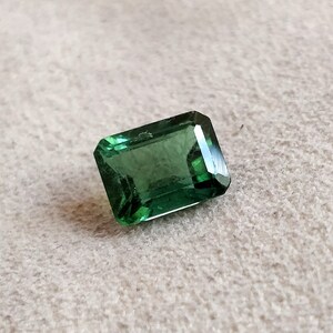 May include: A green emerald cut gemstone on a white background.