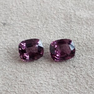 Puede incluir: Dos piedras preciosas de color púrpura talla cojín. Las piedras preciosas son facetadas y tienen un acabado brillante.