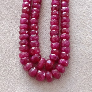 May include: A strand of ruby beads, with a deep red color, is shown in a close-up image. The beads are faceted and have a slightly irregular shape. The beads are strung on a clear thread and are arranged in a loose, flowing pattern.