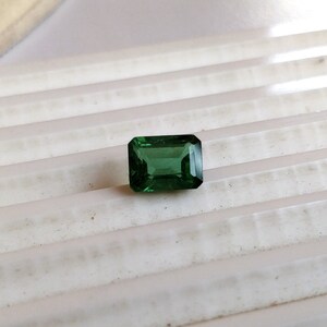 May include: A vibrant green gemstone with a rectangular emerald cut. The gemstone is set against a white background.