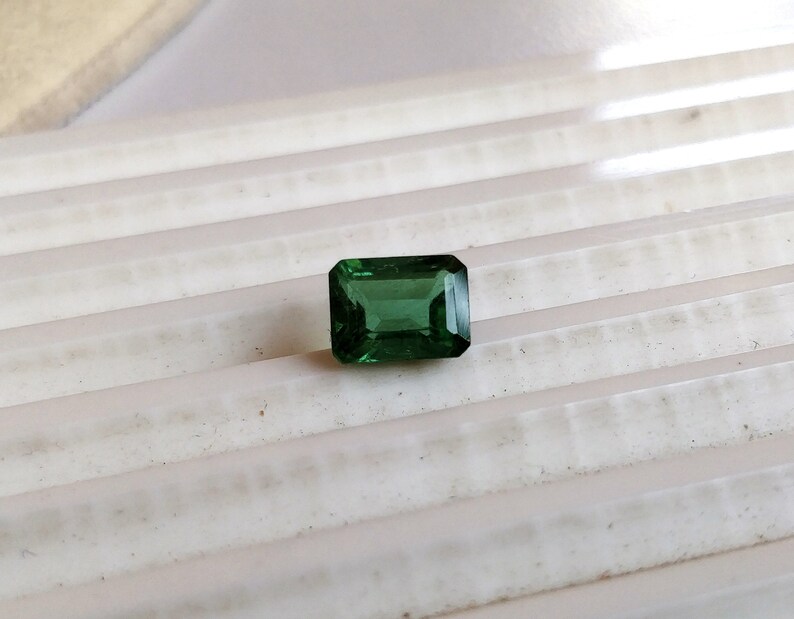 May include: A vibrant green gemstone with a rectangular emerald cut. The gemstone is set against a white background.