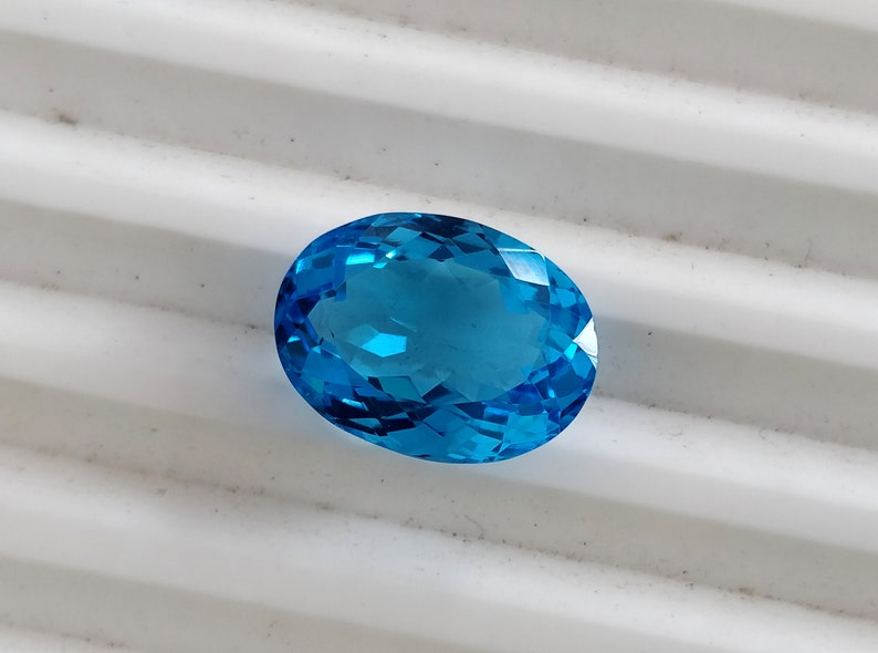 May include: A vibrant blue oval-shaped gemstone with a faceted cut. The gemstone is set against a white background.