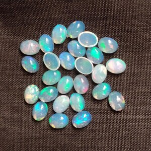 Puede incluir: Un conjunto de ópalos ovalados con apariencia translúcida y lechosa. Los ópalos muestran destellos iridiscentes de azul, verde y rosa, sobre un fondo oscuro. Estas piedras preciosas son ideales para la creación de joyas.