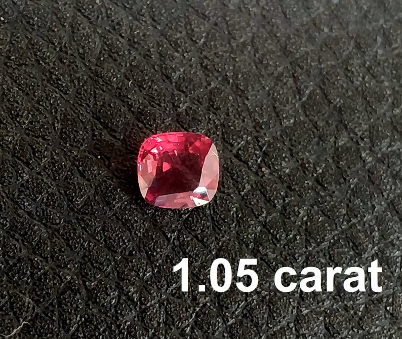 Pode incluir: Uma gema vermelha de 1,05 quilates, provavelmente um rubi, com corte almofada. A gema est&aacute; colocada sobre um fundo preto.