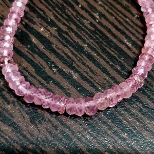 Pode incluir: Um bracelete de pedras preciosas cor-de-rosa feito de pequenas contas facetadas. O bracelete est&aacute; sobre uma superf&iacute;cie de madeira castanho.