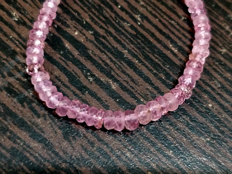 Pode incluir: Um bracelete de pedras preciosas cor-de-rosa feito de pequenas contas facetadas. O bracelete est&aacute; sobre uma superf&iacute;cie de madeira castanho.