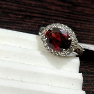 May include: A silver ring with a large oval-shaped red gemstone surrounded by smaller white gemstones.