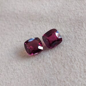 May include: Two deep red square-shaped gemstones on a white background.