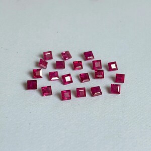 Può includere: Una collezione di venti piccoli rubini a taglio quadrato di un colore rosso intenso e vibrante. Ogni gemma presenta una superficie sfaccettata che riflette la luce, disposta in un gruppo sparso su una superficie neutra e uniforme.