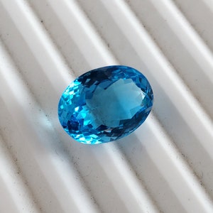 May include: A bright blue oval-shaped gemstone with facets. The gemstone is on a white textured surface.