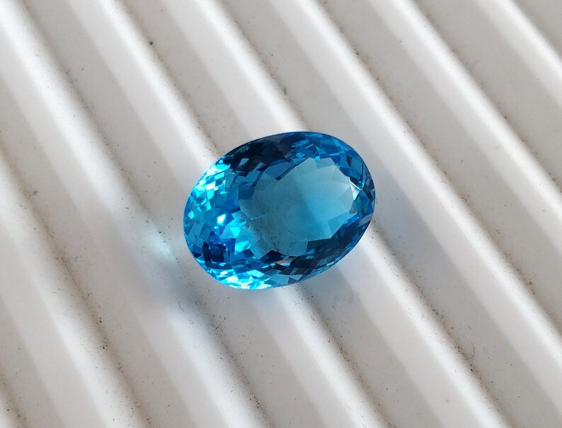 May include: A bright blue oval-shaped gemstone with facets. The gemstone is on a white textured surface.