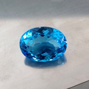 May include: A large, oval-shaped, blue topaz gemstone with a faceted cut. The gemstone is set against a silver background.