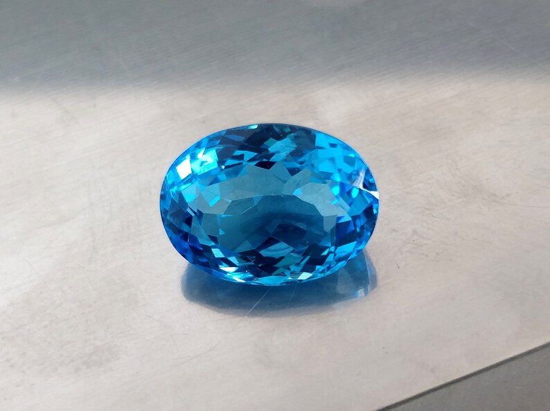 May include: A large, oval-shaped, blue topaz gemstone with a faceted cut. The gemstone is set against a silver background.