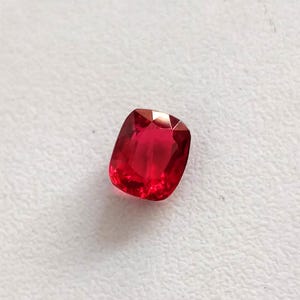 Könnte beinhalten: Ein leuchtend roter Edelstein im Kissenschliff mit einer facettierten Oberfläche, die das Licht reflektiert. Der Stein zeigt einen tiefen, gesättigten karmesinroten Farbton und eine symmetrische, abgerundete quadratische Form mit hoher Klarheit.