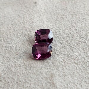 May include: Two purple cushion-cut gemstones on a white textured surface.