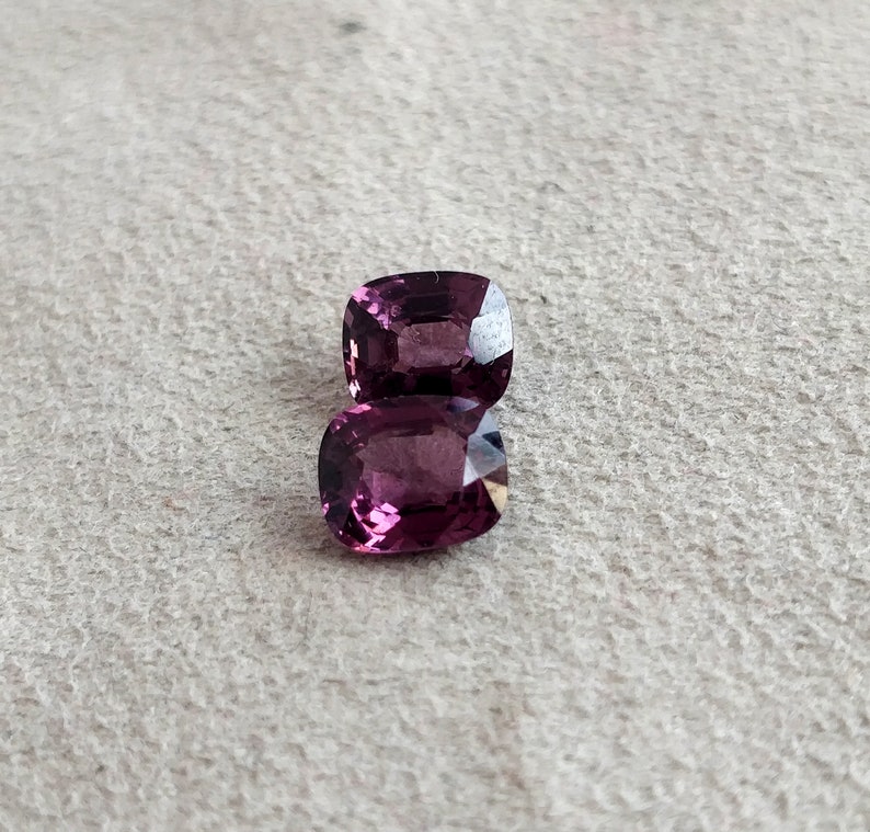 May include: Two purple cushion-cut gemstones on a white textured surface.