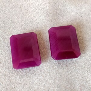 Puede incluir: Dos cabujones de piedra preciosa púrpura, cortados en forma rectangular con bordes biselados. Las piedras preciosas son lisas y pulidas.
