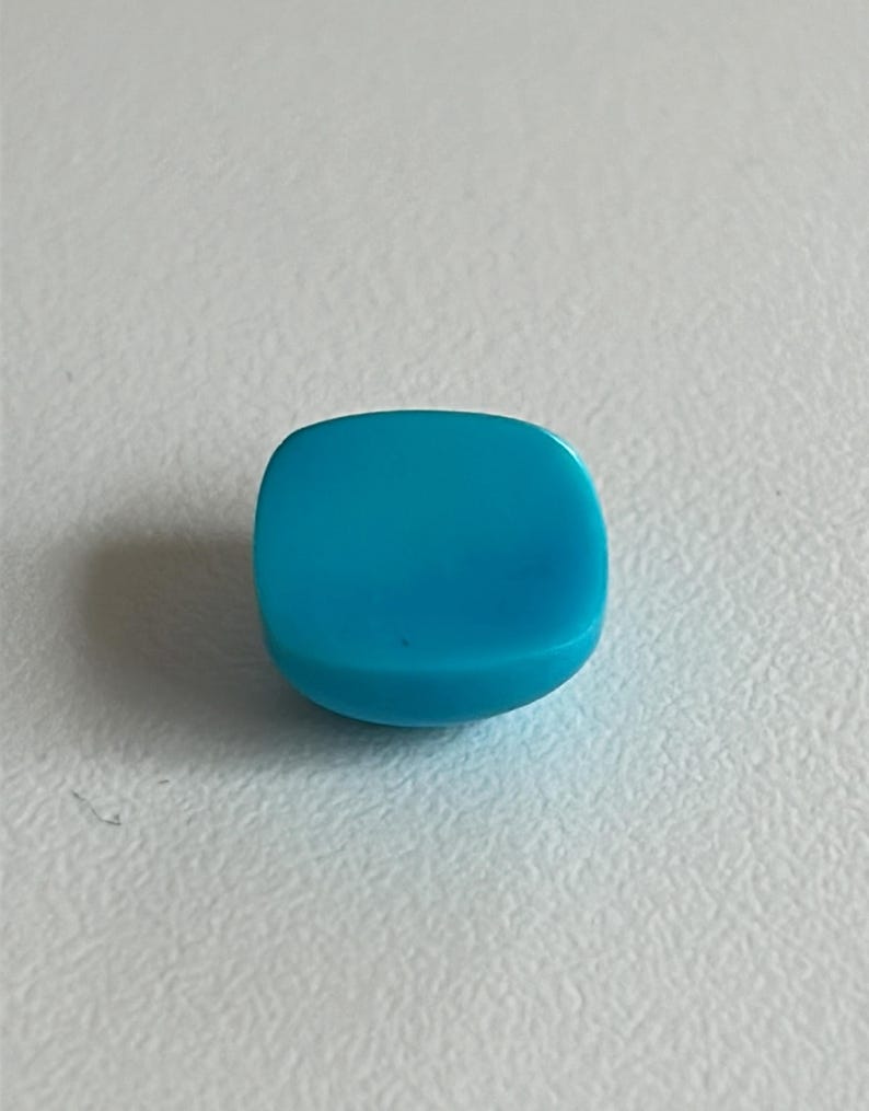 May include: A bright turquoise blue cabochon gemstone, cut in a square shape with rounded edges.