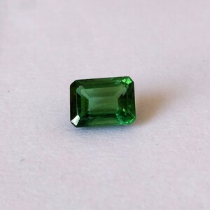 May include: A vibrant green emerald cut gemstone, with a rectangular shape and a polished finish.