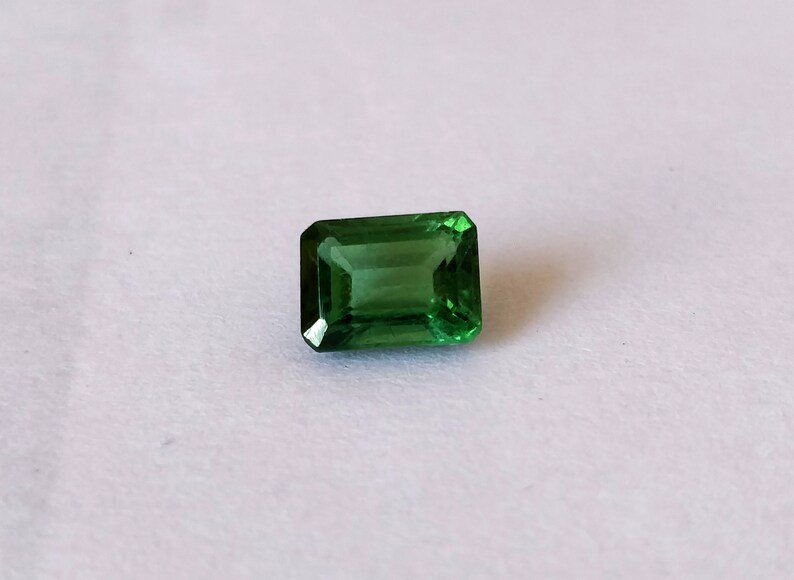 May include: A vibrant green emerald cut gemstone, with a rectangular shape and a polished finish.