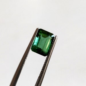 May include: A vibrant green emerald gemstone, cut in an emerald shape, held by tweezers against a white background.