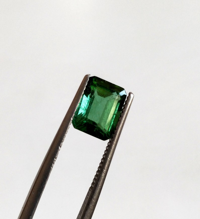 May include: A vibrant green emerald gemstone, cut in an emerald shape, held by tweezers against a white background.