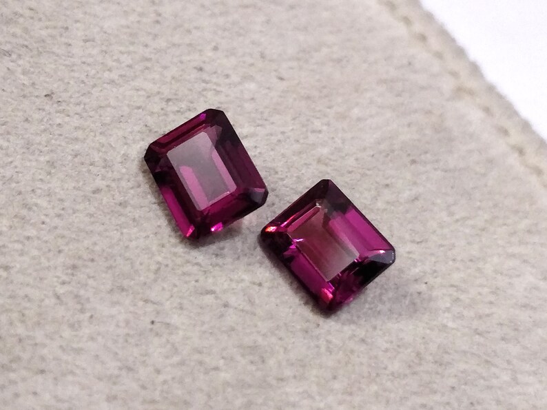 May include: Two purple gemstone rectangular cut loose stones on a white background.