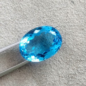 May include: A bright blue oval-shaped gemstone with many facets, held by silver tweezers against a gray textured background.