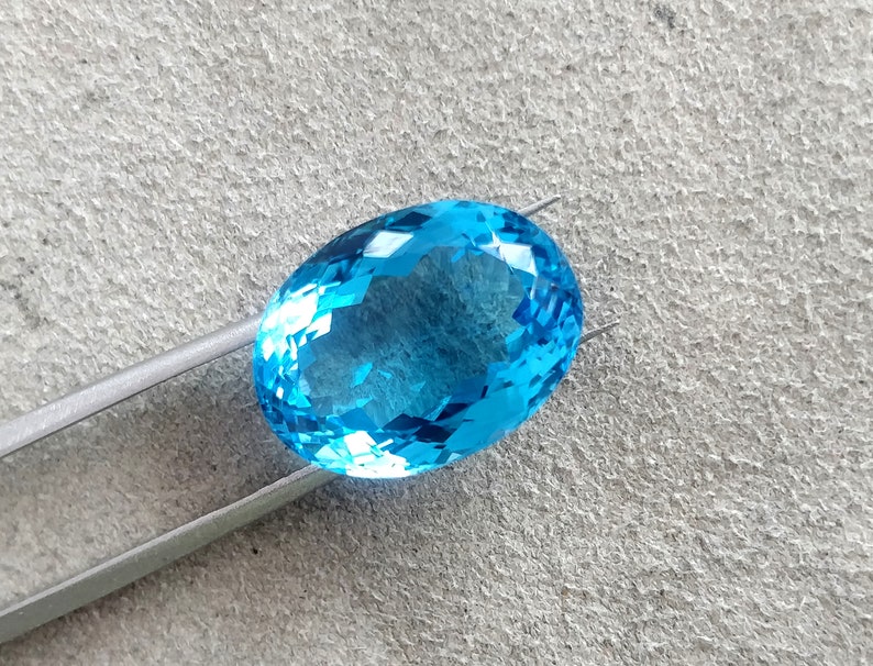 May include: A bright blue oval-shaped gemstone with many facets, held by silver tweezers against a gray textured background.
