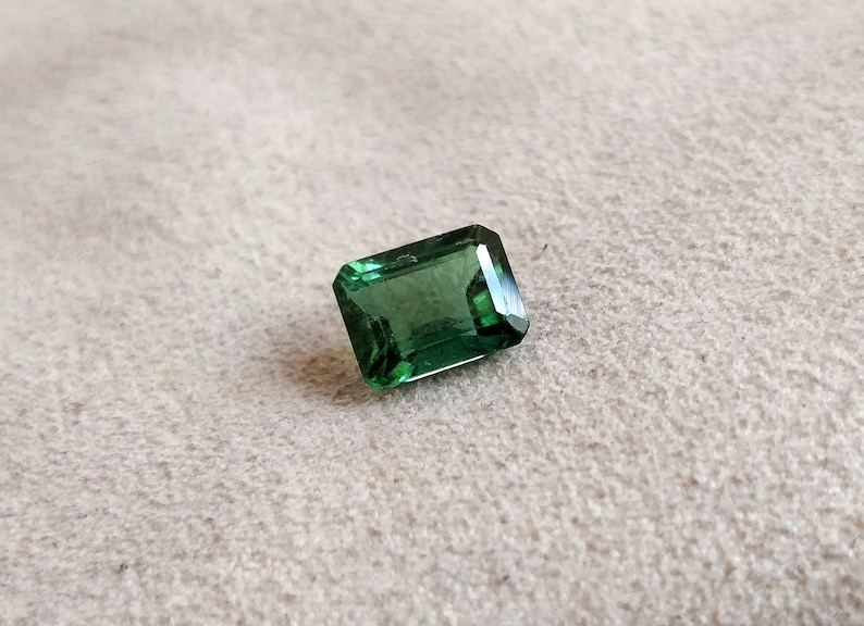 May include: A green emerald cut gemstone on a white background.