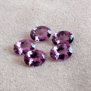 Puede incluir: Cinco piedras preciosas de amatista de forma ovalada en varios tonos de púrpura. Las piedras preciosas están dispuestas en círculo sobre una superficie gris claro.