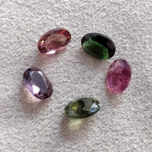 May include: Five oval-shaped gemstones in various colors, including pink, green, and purple. The gemstones are arranged in a circle on a white surface.
