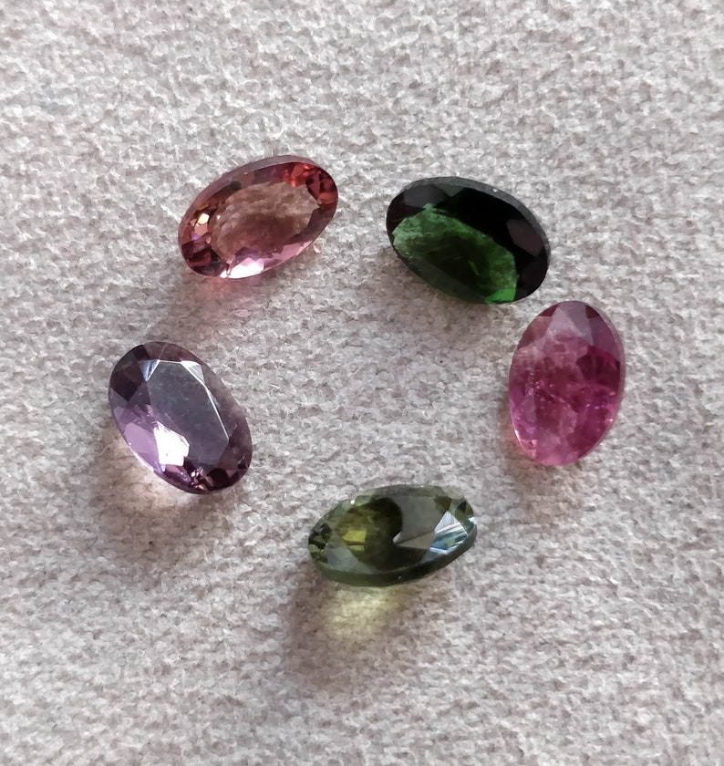 May include: Five oval-shaped gemstones in various colors, including pink, green, and purple. The gemstones are arranged in a circle on a white surface.