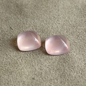 Puede incluir: Dos gemas de cuarzo rosa pulidas, de forma cuadrada. Las piedras tienen un suave tono rosa translúcido y una superficie lisa y redondeada. Se muestran sobre un fondo texturizado de tono neutro.