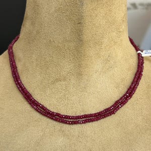 May include: A double-strand necklace featuring small, faceted, deep red gemstones. The necklace is displayed on a beige mannequin bust, showcasing the jewelry's color and design. The gemstones appear to be evenly sized and closely strung.