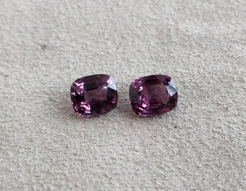 May include: Two cushion-cut, purple gemstones. The gemstones are faceted and have a glossy finish.