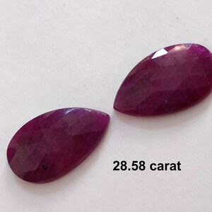 Puede incluir: Dos piedras preciosas talladas en forma de pera, cada una de un color rojo púrpura profundo y rico. Las piedras preciosas se muestran sobre una superficie blanca, con el texto "28,58 quilates" debajo.