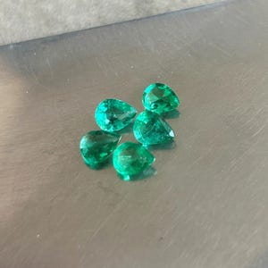 K&ouml;nnte beinhalten: F&uuml;nf lose, birnenf&ouml;rmige Smaragd-Edelsteine mit leuchtend gr&uuml;ner Farbe und facettierten Oberfl&auml;chen. Die Steine sind in einer kleinen Gruppe angeordnet und zeigen ihre durchscheinende Qualit&auml;t und ihre tropfenf&ouml;rmigen Silhouetten.
