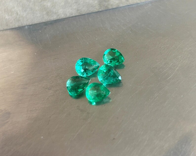 K&ouml;nnte beinhalten: F&uuml;nf lose, birnenf&ouml;rmige Smaragd-Edelsteine mit leuchtend gr&uuml;ner Farbe und facettierten Oberfl&auml;chen. Die Steine sind in einer kleinen Gruppe angeordnet und zeigen ihre durchscheinende Qualit&auml;t und ihre tropfenf&ouml;rmigen Silhouetten.