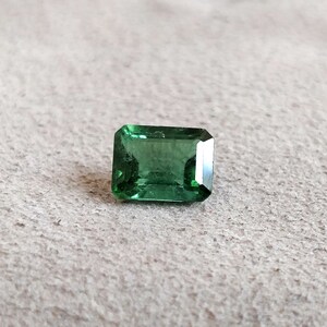 May include: A green emerald cut gemstone on a white textured surface.