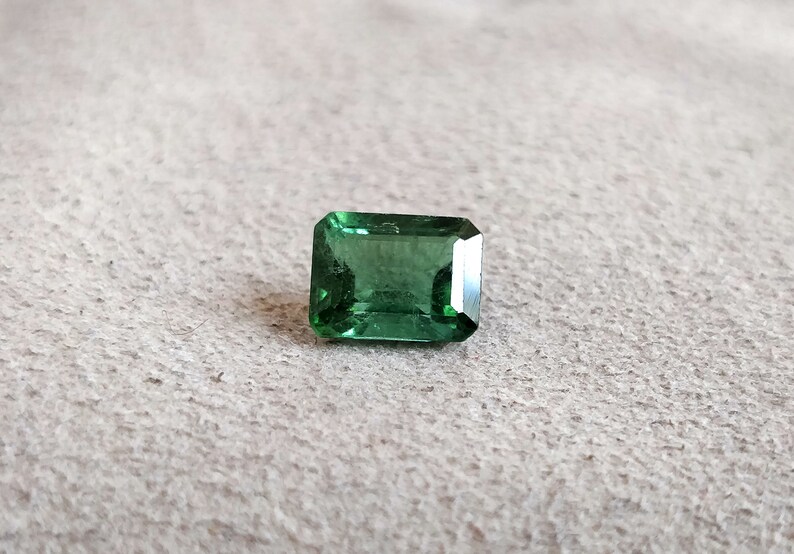 May include: A green emerald cut gemstone on a white textured surface.
