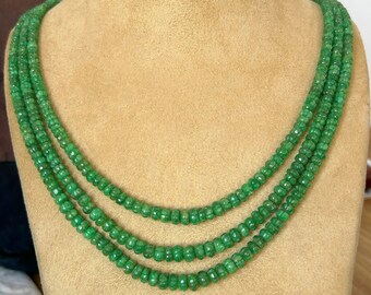 May include: A three-strand emerald green gemstone necklace. The necklace is made of small, round beads and is displayed on a brown velvet mannequin.