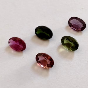 May include: Five oval-shaped gemstones in various colors, including pink, green, and purple. The gemstones are arranged on a white surface.